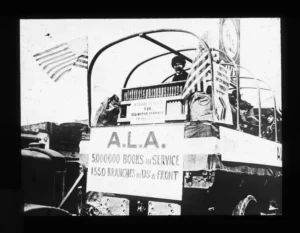 A black and white photo of a military truck loaded with books and American flags. A sign on the back reads, "A.L.A. 5000000 BOOKS IN SERVICE, 1550 BRANCHES IN U.S. & FRONT." A soldier smiles from inside the truck.