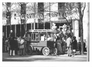 A lively crowd gathered in front of a building decorated with balloons and a "GROUND BREAKING" banner. A vintage-style popcorn cart sits in the foreground. People appear to be celebrating a new opening.