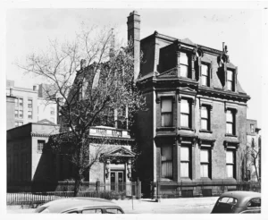 A black and white photo of a grand, multi-story mansion with a steep roof and intricate window moldings. A few cars are parked outside, and a tree without leaves stands in the front yard.