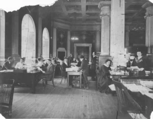 A vintage photograph of a large room with ornate columns and high ceilings. Men and women are seen working at desks, reading, writing, and typing, capturing the bustling activity of a historical library office.
