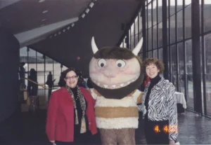 Two smiling women standing beside a person in a large costume of a monster character from “Where the Wild Things Are,” inside a modern building with floor-to-ceiling windows.