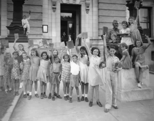 A group of children proudly holding up books in front of a library building. Some stand beside or on a statue of a seated man. The children are smiling and appear to be celebrating reading