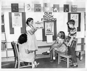 Four children in a classroom setting present favorite books in front of a display board labeled “OUR BOOK FRIENDS.” One girl holds up a drawing of a book titled "Peanuts the Pony."