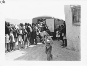 Children gathered in line near a bookmobile parked beside a building. Some hold books, while a boy runs past the camera holding multiple items. The background shows open landscape and mountains.