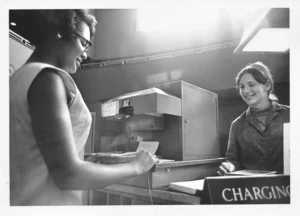 A black and white photo shows a young African American woman in glasses smiling as she checks out a book at a library counter. A librarian with long hair smiles back at her from behind the counter, next to a book charging machine.
