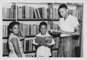 Three African American children are shown in a library. A girl on the left smiles at the camera, a boy in the center reads a book, and an older boy on the right also reads a book. Bookshelves are filled with books