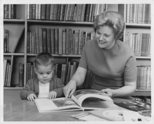 A smiling blonde woman, Dorothy Anderson, points to a large open book while sitting at a table with a young child, who is looking intently at the book. Bookshelves filled with books are in the background.