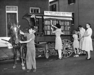A black and white photo shows a horse-drawn "Pratt Library's Book Wagon" with bookshelves, from which a woman is selecting a book. A boy holds the horse's bridle, and several women and children are gathered around.