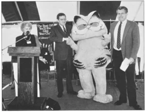 The cartoon character Garfield in a mascot costume stands between two men in suits, while a woman speaks at a podium to the left. A sign behind them reads "The Best Gift You'll Ever Give Your Child: A Library Card."
