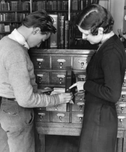 A black and white photo shows a young man and woman looking at a card catalog. The man is pulling out a card, while the woman points to a book. Bookshelves are visible in the background.