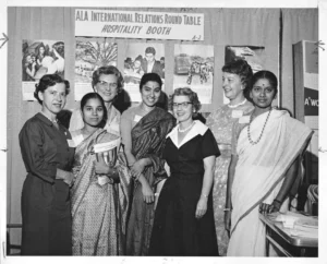 Eight women, some dressed in saris, are posing together in front of a backdrop with the text "ALA International Relations Round Table Hospitality Booth" and several images.
