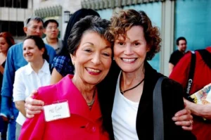 Two smiling women, both named Judy (Judy Blume on the right with curly hair and a dark jacket, and Judy King on the left with short dark hair and a pink jacket), stand close together with their arms around each other, looking at the camera. Other people are blurred in the background.