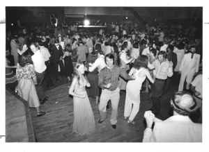 A black and white photo captures a lively disco scene with many people dancing on a wooden floor in a large room.