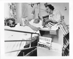A black and white photo shows a librarian, wearing glasses and a white uniform, holding a book for a male patient who is lying in a hospital bed and also holding a book. A book cart is beside the bed.