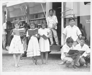 A black and white photo shows a group of barefoot children, primarily of Asian descent, reading books outdoors near a large bookmobile with its side shelves open.