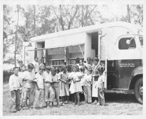 A black and white photo shows a group of children gathered around a bookmobile in a grassy outdoor setting, many holding books. A woman is standing near the open side of the bookmobile.