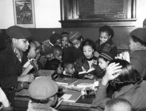 A black and white photo shows a group of diverse children, many wearing hats, gathered around a woman who is reading a book aloud at a table in what appears to be a library.