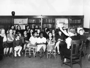 A black and white photo shows a diverse group of children sitting on benches and chairs in front of bookshelves, attentively watching a woman who is holding up a book and reading to them.