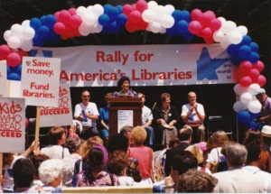 A color photo captures a "Rally for America's Libraries" event outdoors, featuring a stage with a speaker at a podium under a red, white, and blue balloon arch. Signs in the crowd advocate for library funding.