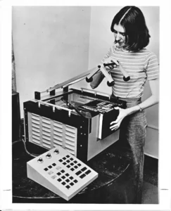 A black and white photo shows a young woman operating a large, boxy machine with a book placed on its surface and a separate control panel on the table. This is identified as a Kurzweil Reading Machine.