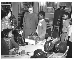 A black and white photo shows a group of elementary school-aged children, some wearing headphones, gathered around a large reel-to-reel tape recorder on a table. Two adult women are standing with them, observing.