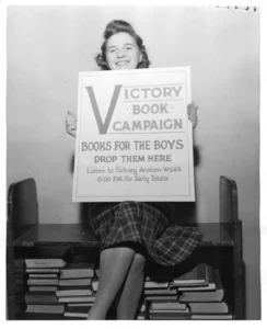 A black and white photo shows a young woman with a smiling expression, seated on a wooden shelf filled with stacks of books. She is holding up a sign that reads "Victory Book Campaign - BOOKS FOR THE BOYS - DROP THEM HERE - Listen to Sidney Andorn - WGAR - 6:00 P.M. For Daily Totals."