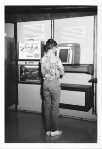 A black and white photo shows a young person with short hair and glasses, wearing a plaid shirt and slacks, standing with their back to the camera. They are facing a display with early electronic equipment, including a monitor and a device with exposed wiring, and informational text on the wall above.