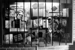 A barred glass display window featuring banned books and posters about censorship. The central sign promotes Banned Books Week (Sept 26 - Oct 3), with slogans like "Celebrate the Freedom to Read."