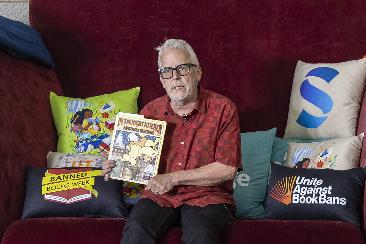 A man sits on a very large chair holding up a copy of children's book "In the Night Kitchen" by Maurice Sendak